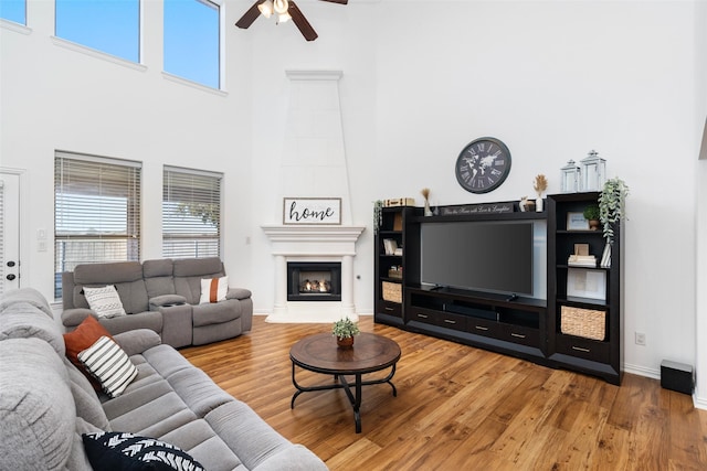 living room featuring ceiling fan, wood finished floors, a warm lit fireplace, a towering ceiling, and baseboards