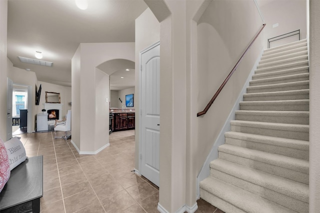 stairs featuring a warm lit fireplace, visible vents, baseboards, arched walkways, and tile patterned floors