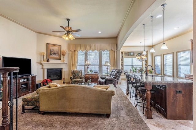 living room featuring a glass covered fireplace, crown molding, arched walkways, and ceiling fan with notable chandelier