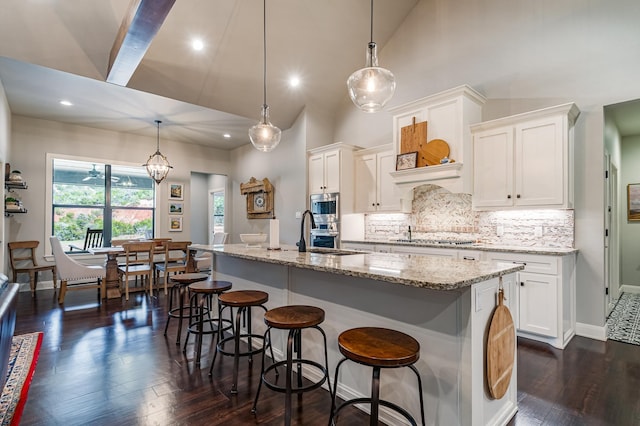 kitchen featuring a kitchen island with sink, white cabinetry, backsplash, light stone countertops, and dark wood-style floors