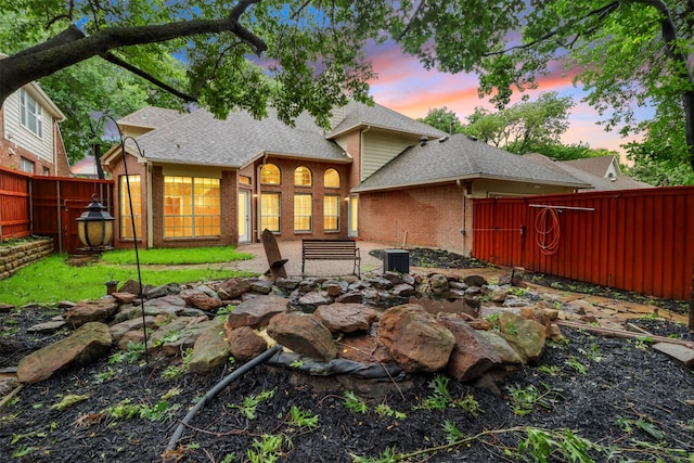 back of property at dusk featuring brick siding, a shingled roof, and a fenced backyard