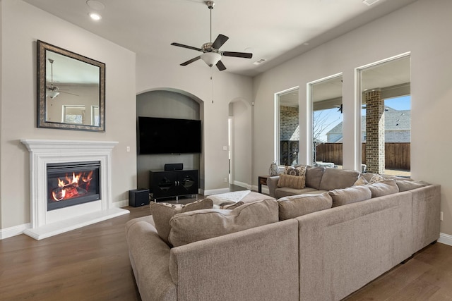 living area featuring ceiling fan, baseboards, dark wood-style flooring, and a glass covered fireplace