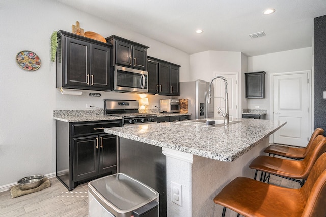 kitchen with a center island with sink, dark cabinets, stainless steel appliances, a kitchen bar, and a sink