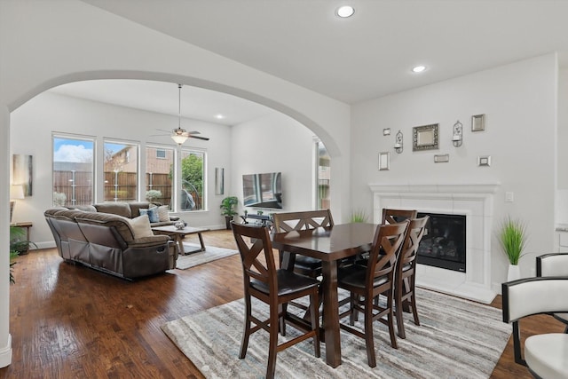 dining space featuring baseboards, arched walkways, a tiled fireplace, wood finished floors, and recessed lighting