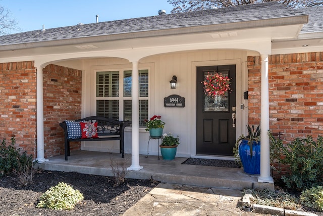 doorway to property featuring covered porch, brick siding, and roof with shingles