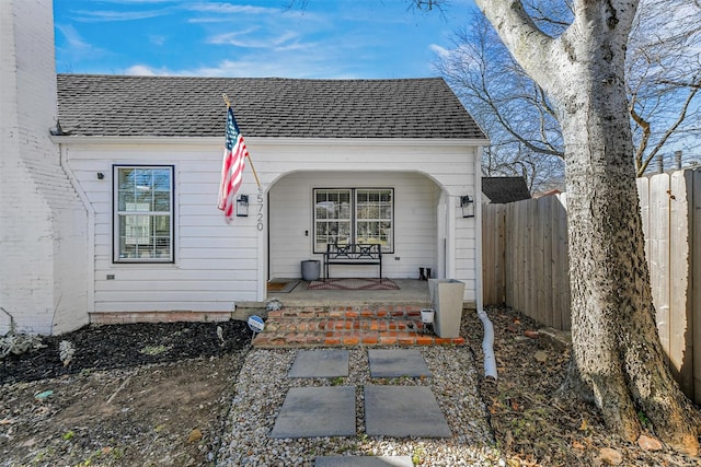 doorway to property featuring covered porch, a shingled roof, and fence