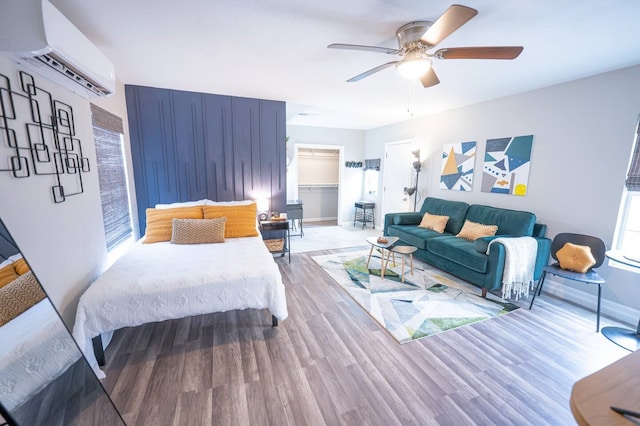 bedroom featuring a walk in closet, a wall unit AC, ceiling fan, wood finished floors, and baseboards