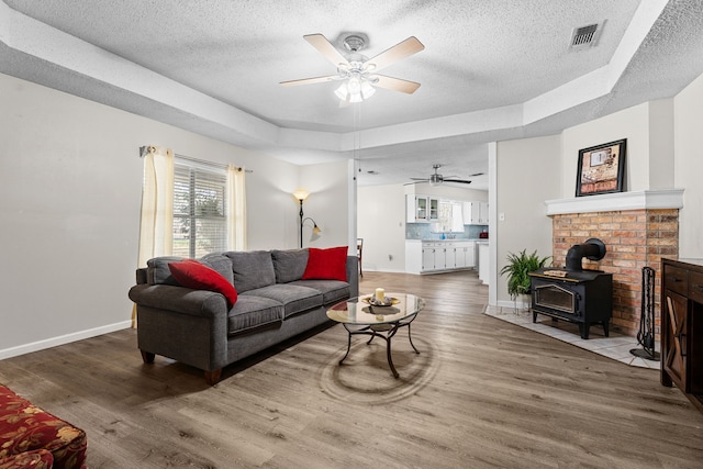 living area with a tray ceiling, visible vents, a wood stove, wood finished floors, and baseboards