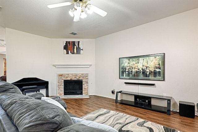 living room featuring baseboards, visible vents, a ceiling fan, wood finished floors, and a stone fireplace