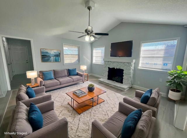 living room featuring vaulted ceiling, a stone fireplace, a textured ceiling, ceiling fan, and baseboards