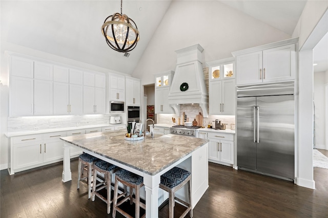 kitchen with dark wood-type flooring, premium range hood, built in appliances, a kitchen breakfast bar, and white cabinetry