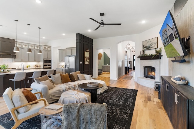 living room with ceiling fan, arched walkways, a brick fireplace, light wood-style floors, and recessed lighting