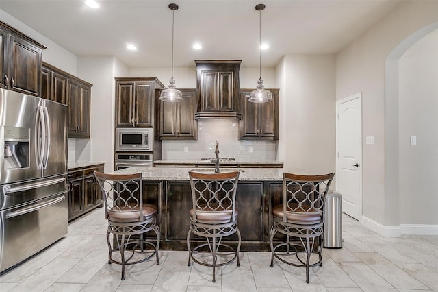 kitchen with light stone counters, arched walkways, stainless steel appliances, an island with sink, and a kitchen bar