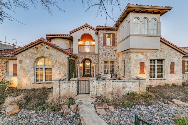 mediterranean / spanish house featuring a balcony, a tiled roof, stone siding, a gate, and stucco siding