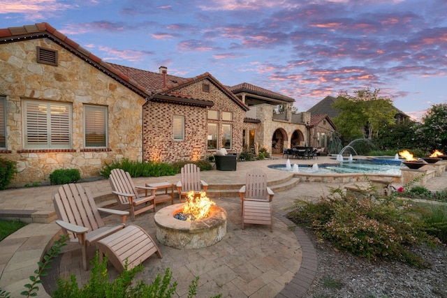 back of house at dusk with a fire pit, a tiled roof, stone siding, an outdoor pool, and a patio area