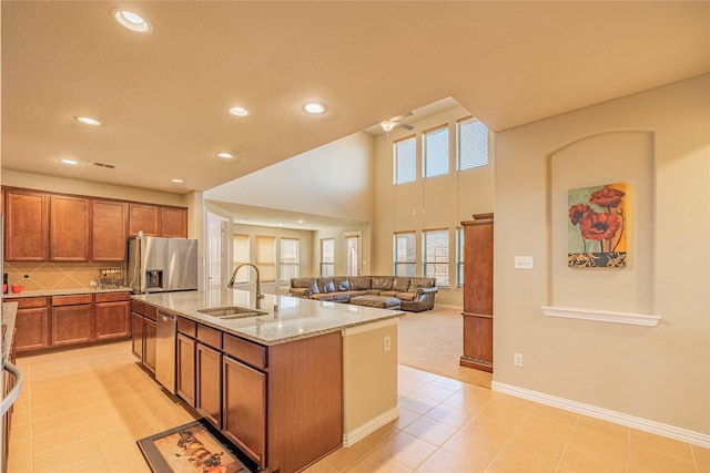 kitchen featuring a center island with sink, brown cabinetry, light stone counters, stainless steel appliances, and a sink