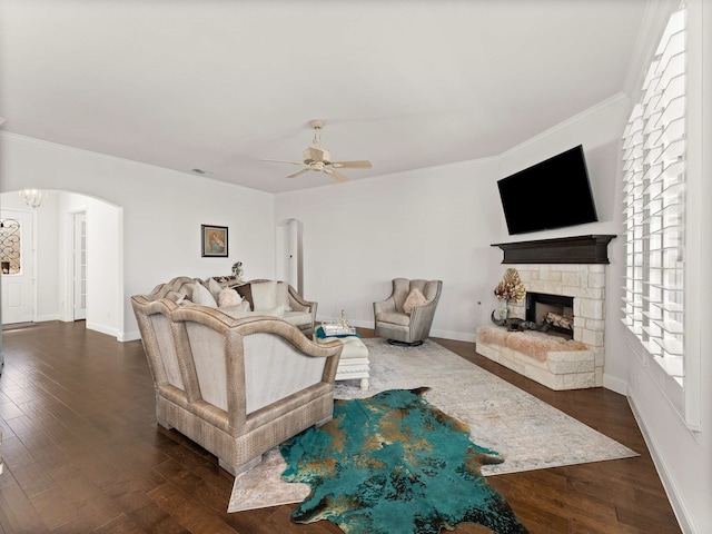 living room featuring arched walkways, dark wood-type flooring, crown molding, a stone fireplace, and ceiling fan with notable chandelier