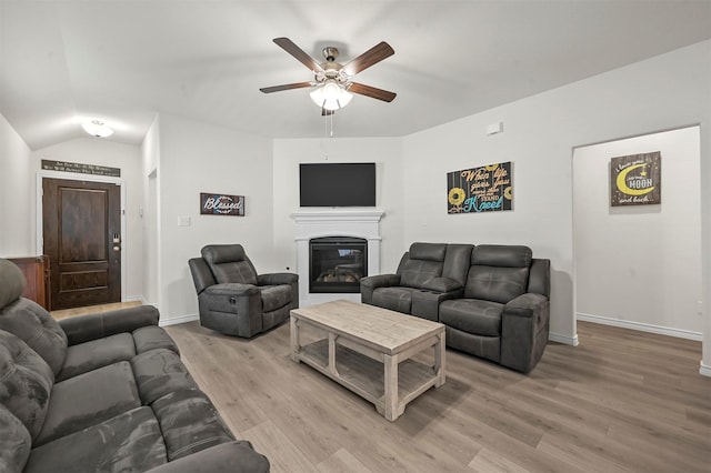 living area featuring baseboards, a ceiling fan, a glass covered fireplace, lofted ceiling, and light wood-style floors