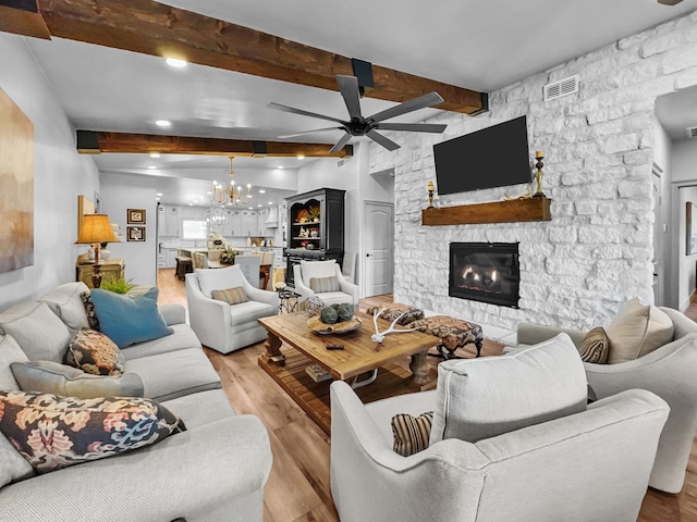 living room featuring a stone fireplace, ceiling fan with notable chandelier, visible vents, light wood-style floors, and beam ceiling