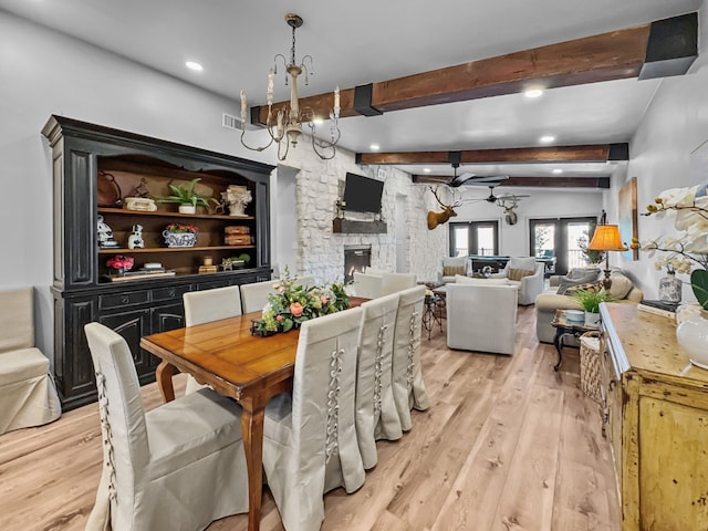 dining room with a stone fireplace, light wood-style flooring, ceiling fan with notable chandelier, visible vents, and beamed ceiling