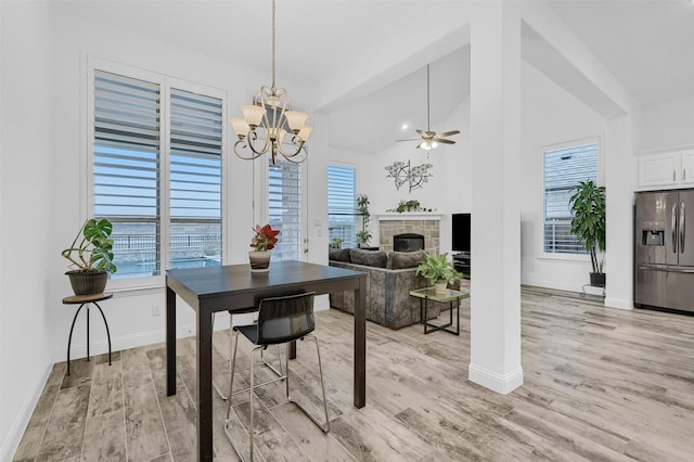 dining area with baseboards, ceiling fan with notable chandelier, a fireplace, light wood-style floors, and high vaulted ceiling