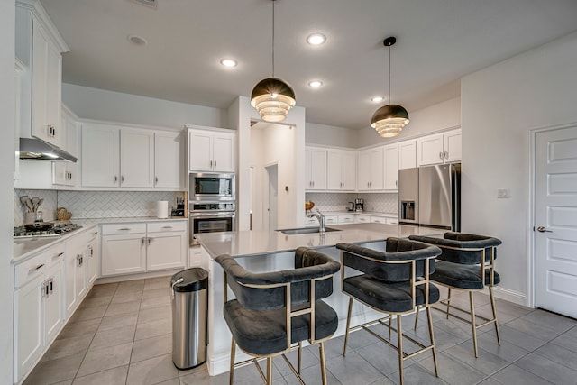 kitchen with a breakfast bar area, under cabinet range hood, a sink, appliances with stainless steel finishes, and an island with sink