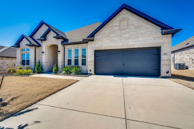 french provincial home with driveway, a shingled roof, central AC unit, an attached garage, and brick siding