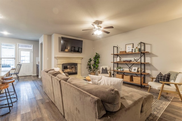 living area featuring a glass covered fireplace, dark wood-style flooring, ceiling fan, and baseboards