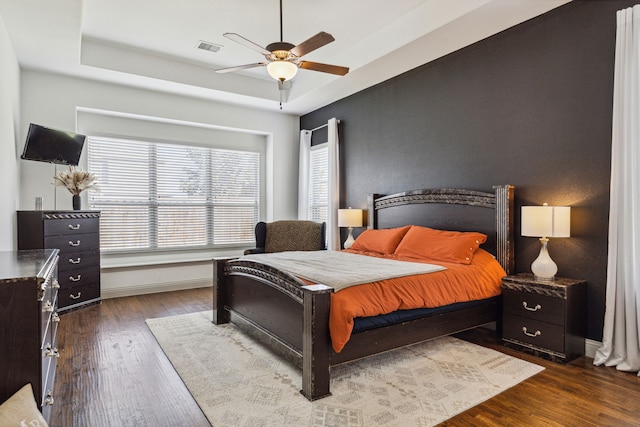 bedroom featuring a ceiling fan, visible vents, baseboards, dark wood-style floors, and a tray ceiling