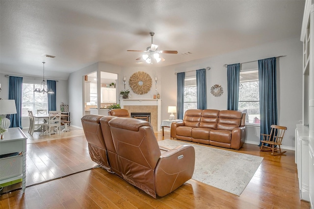 living room with a tile fireplace, ceiling fan with notable chandelier, visible vents, baseboards, and light wood-type flooring