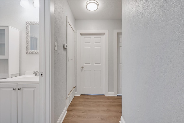 corridor featuring a textured wall, a sink, light wood-style flooring, and baseboards