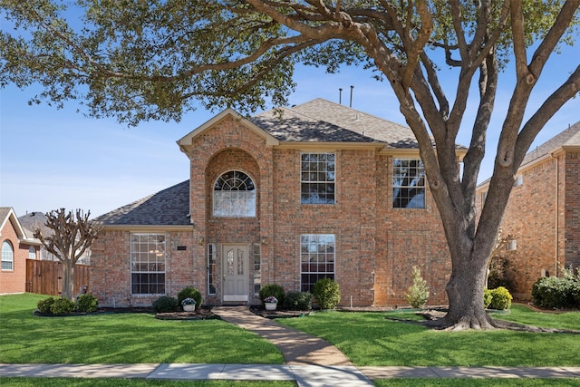 traditional home featuring roof with shingles, fence, a front lawn, and brick siding