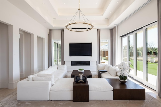 living room with baseboards, a raised ceiling, a glass covered fireplace, a high ceiling, and a notable chandelier