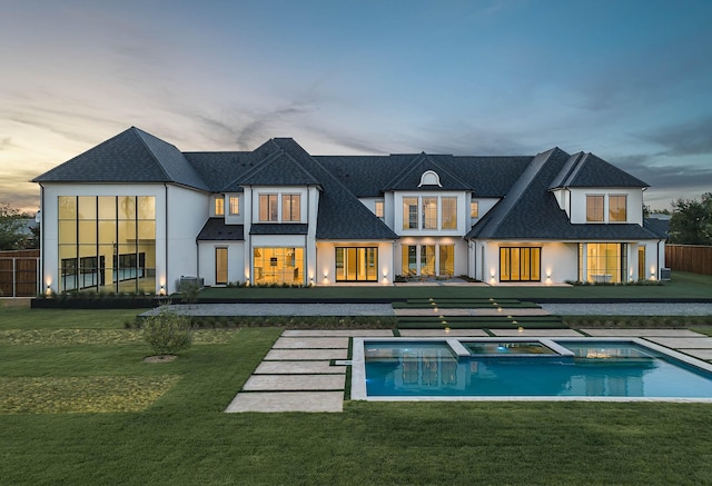 back of house at dusk with a shingled roof, a pool with connected hot tub, and a yard