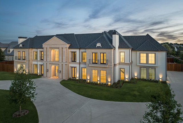 back of property at dusk with fence, stucco siding, a chimney, concrete driveway, and a lawn