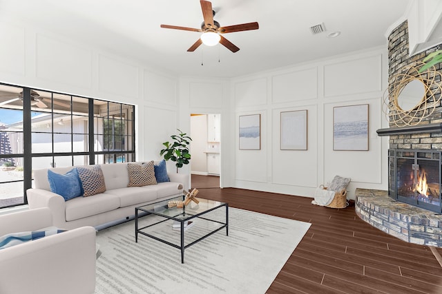 living room with visible vents, dark wood finished floors, a ceiling fan, a brick fireplace, and a decorative wall