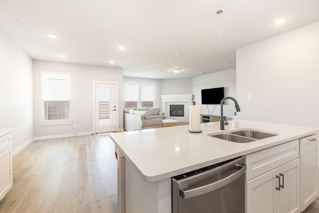 kitchen featuring a sink, stainless steel dishwasher, light wood finished floors, an island with sink, and a glass covered fireplace