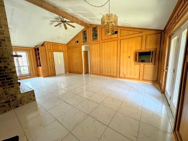 unfurnished living room featuring marble finish floor, wood walls, lofted ceiling with beams, and ceiling fan with notable chandelier