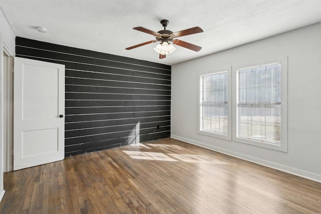 empty room with ceiling fan, wood walls, a textured ceiling, baseboards, and hardwood / wood-style flooring