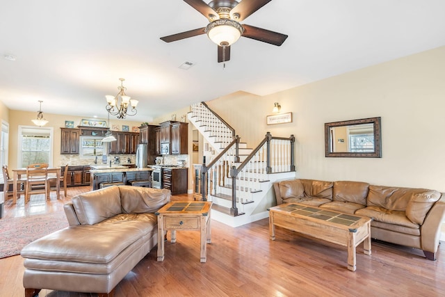 living area featuring stairs, light wood finished floors, ceiling fan with notable chandelier, and visible vents