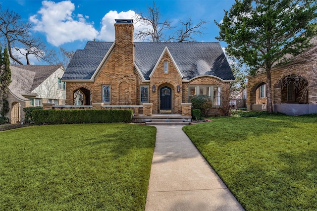 english style home featuring brick siding, a front lawn, a chimney, and a shingled roof