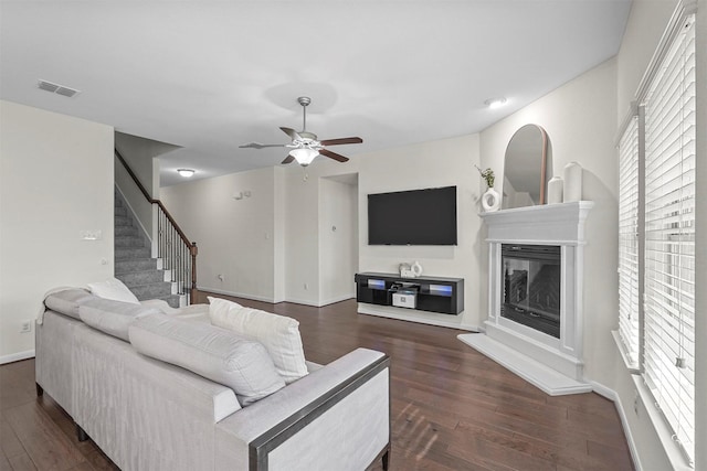 living room featuring ceiling fan, dark wood-type flooring, visible vents, stairway, and a glass covered fireplace