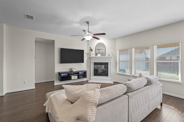 living room featuring dark wood-style floors, a glass covered fireplace, visible vents, and ceiling fan