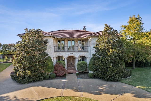 mediterranean / spanish home featuring a balcony, stone siding, a tile roof, a chimney, and a front yard