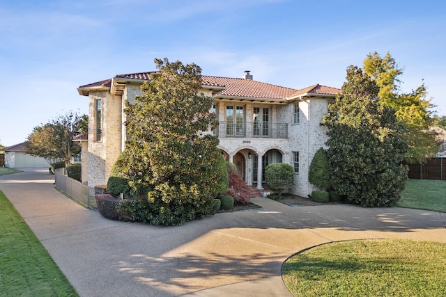 mediterranean / spanish-style house featuring a balcony, stone siding, a tile roof, and fence