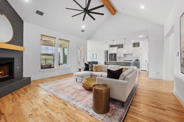 living room with high vaulted ceiling, a fireplace, a ceiling fan, beam ceiling, and light wood finished floors