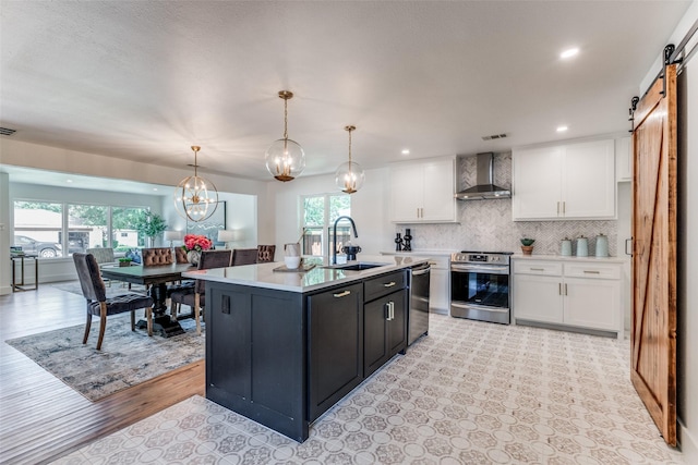 kitchen with a barn door, appliances with stainless steel finishes, wall chimney range hood, white cabinetry, and a sink