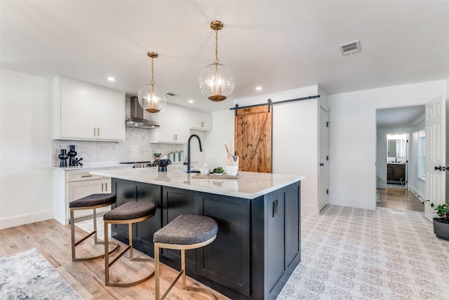 kitchen with a barn door, visible vents, white cabinets, wall chimney exhaust hood, and tasteful backsplash