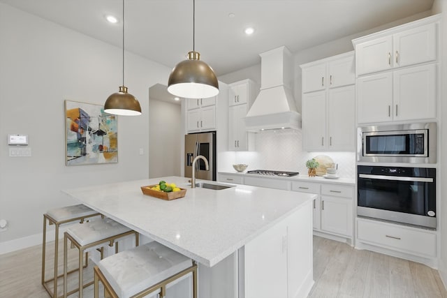 kitchen with custom range hood, decorative backsplash, appliances with stainless steel finishes, a sink, and an island with sink