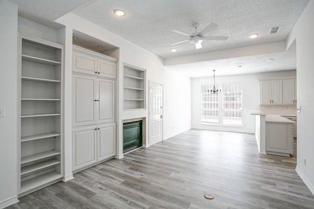 unfurnished living room featuring light wood finished floors, visible vents, a glass covered fireplace, a textured ceiling, and ceiling fan with notable chandelier